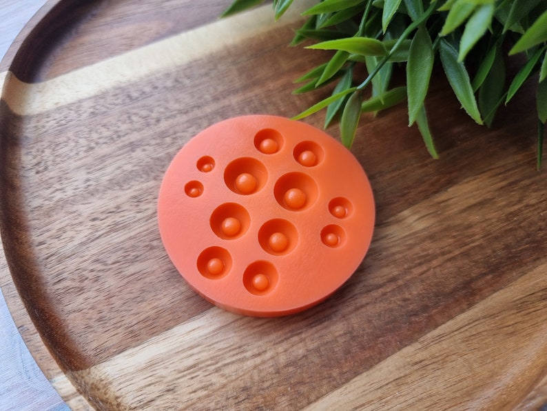 K&ouml;nnte beinhalten: Orangefarbene Silikonform mit 10 runden Vertiefungen in verschiedenen Gr&ouml;&szlig;en. Die Form befindet sich auf einer Holzoberfl&auml;che.