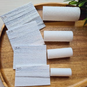 Könnte beinhalten: Eine Auswahl an weißen Schleifpads und zylindrischen Schleifhülsen, angeordnet auf einem Holztablett. Die quadratischen Pads sind strukturiert und mit der Zahl "10" bedruckt. Die Hülsen sind weiß, strukturiert und zylindrisch.