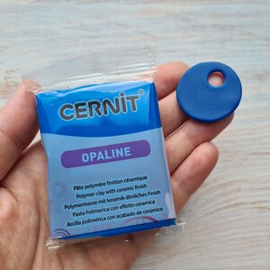 Może przedstawiać: Opakowanie niebieskiej gliny polimerowej Cernit Opaline z ceramicznym wykończeniem oraz osobna niebieska zawieszka z otworem. Opakowanie zawiera tekst w wielu językach. Przedmioty leżą na jasnej drewnianej powierzchni.