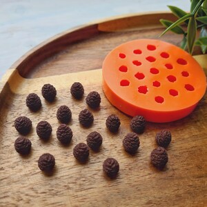 Pode incluir: Um close-up de uma bandeja de madeira com uma forma de silicone para fazer pequenas formas de pinhas. A forma &eacute; laranja e tem 24 cavidades individuais. H&aacute; v&aacute;rias pequenas formas de pinhas marrons espalhadas ao redor da forma na bandeja.