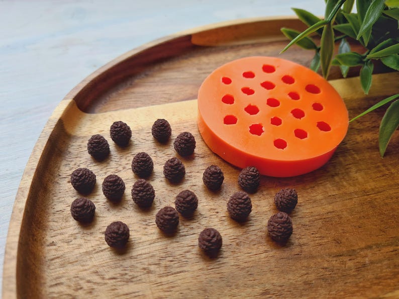 Pode incluir: Um close-up de uma bandeja de madeira com uma forma de silicone para fazer pequenas formas de pinhas. A forma &eacute; laranja e tem 24 cavidades individuais. H&aacute; v&aacute;rias pequenas formas de pinhas marrons espalhadas ao redor da forma na bandeja.