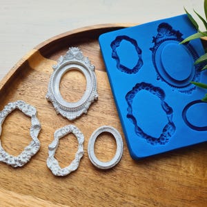 May include: A blue silicone mold with various oval and round frame designs, alongside several cast white resin frames. The frames are ornate, with detailed edges. The items are arranged on a wooden tray, with greenery in the background.