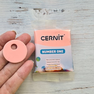 May include: A pink package of polymer clay labeled "CERNIT NUMBER ONE" with the text "Pâte polymère opaque Opaque polymer clay Opake Polymermasse Pasta Polimerica oраса Arcilla polimerica opaco" in multiple languages. A pink circle of polymer clay with a hole in the center is in the foreground.