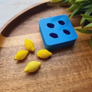 Peut inclure: Un moule en silicone bleu avec six cavités rondes, parfait pour faire de petites friandises en forme de citron. Quatre bonbons jaunes en forme de citron sont sur une surface en bois à côté du moule.