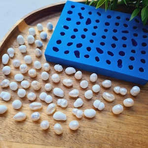 Op de afbeelding: Een rechthoekige blauwe siliconen mal met een raster van kleine holtes ligt naast een verzameling van talrijke losse, onregelmatige witte en gebroken witte parels. De items liggen op een rond houten oppervlak met een zichtbare houtnerf.