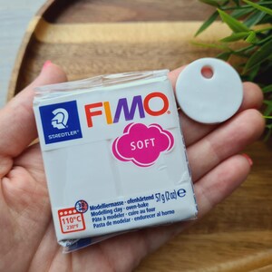May include: A package of white FIMO Soft modeling clay, 2 oz, with a pink cloud-shaped label. A white, round, flat tag with a hole is next to the package. The package has the Staedtler logo and oven-bake instructions.