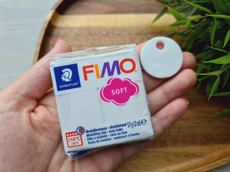May include: A package of white FIMO Soft modeling clay, 2 oz, with a pink cloud-shaped label. A white, round, flat tag with a hole is next to the package. The package has the Staedtler logo and oven-bake instructions.