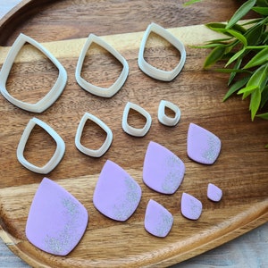 May include: A set of eight white plastic teardrop-shaped cookie cutters and six purple teardrop-shaped fondant pieces with silver glitter on a wooden surface.