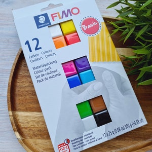 May include: A pack of 12 colours of Fimo Basic modelling clay. The pack is white with a red circle that says "Basic" and shows a close-up of a hand working with the clay. The clay comes in a variety of colours, including white, orange, yellow, red, pink, purple, blue, green, brown, and black.