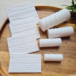 Op de afbeelding: Witte, getextureerde onderzetters en cilindrische objecten zijn gerangschikt op een houten dienblad. De onderzetters hebben een houtnerfpatroon en kleine gaatjes. De cilindrische objecten hebben een geribbeld ontwerp. De dienblad heeft een warme bruine kleur.
