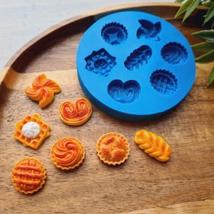 Op de afbeelding: Een blauwe siliconen mal met diverse gebaksvormen, samen met diverse miniatuurgebakjes in oranje en geel. De gebakjes omvatten een wafel, taart en croissant, allemaal gerangschikt op een houten dienblad. De mal heeft vormen als een vlinder en een hart.