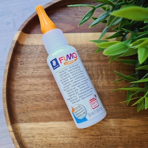 May include: A clear plastic bottle of FIMO Liquid, a translucent gel for oven-hardening polymer clay. The bottle is labeled with instructions in multiple languages and has a 130°C temperature rating. The bottle is sitting on a wooden surface.