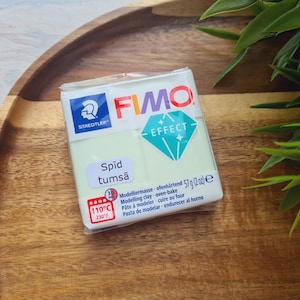 May include: A package of FIMO Effect modeling clay, 57g (2 oz), in a light green color. The package features the Staedtler logo and the text "Spīd tumsā" and "Effect". The clay is oven-bake and is on a wooden tray.