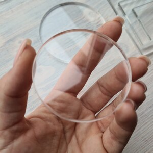 May include: A clear round glass disc held in a hand. The disc is thick and has a smooth surface.