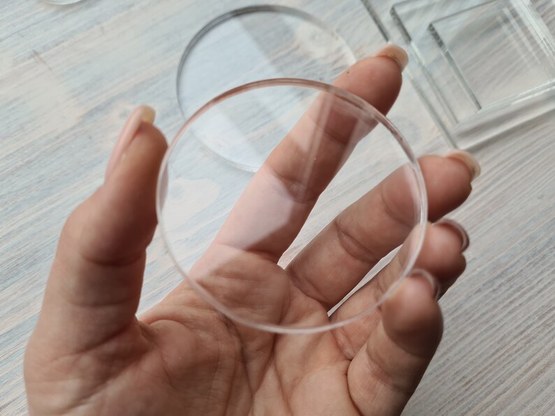 May include: A clear round glass disc held in a hand. The disc is thick and has a smooth surface.