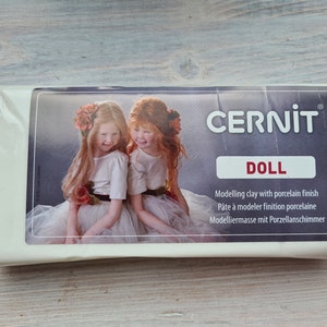 Puede incluir: Un paquete de arcilla de modelado Cernit Doll con acabado de porcelana. La etiqueta presenta una imagen de dos ni&ntilde;as. El texto de la etiqueta incluye "DOLL" y la descripci&oacute;n de la arcilla en varios idiomas.