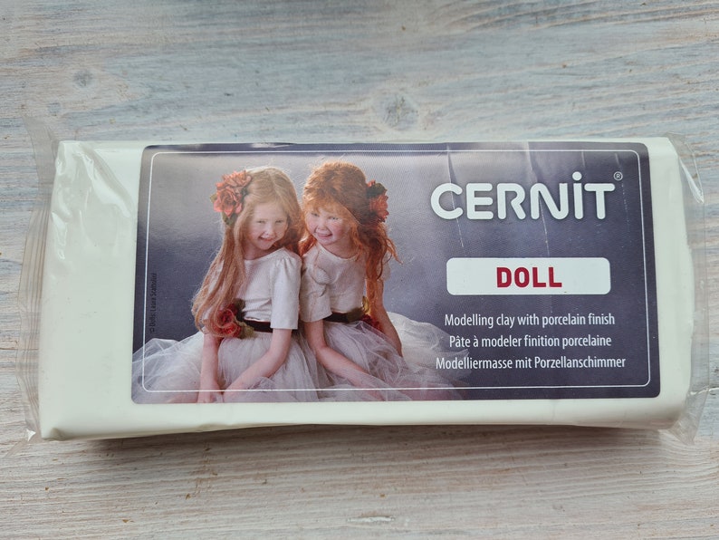 Puede incluir: Un paquete de arcilla de modelado Cernit Doll con acabado de porcelana. La etiqueta presenta una imagen de dos ni&ntilde;as. El texto de la etiqueta incluye "DOLL" y la descripci&oacute;n de la arcilla en varios idiomas.