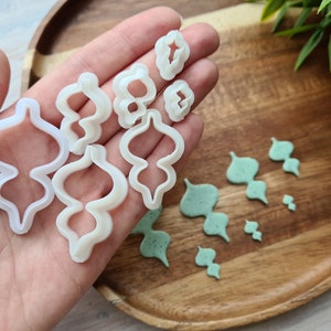 Puede incluir: Un juego de cortadores de galletas de pl&aacute;stico blanco en varias formas, incluyendo una l&aacute;grima, un remolino y una hoja. Los cortadores est&aacute;n sobre una superficie de madera con recortes de fondant verde.