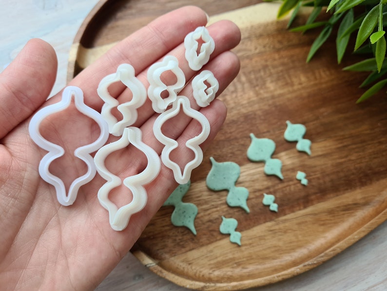 Puede incluir: Un juego de cortadores de galletas de pl&aacute;stico blanco en varias formas, incluyendo una l&aacute;grima, un remolino y una hoja. Los cortadores est&aacute;n sobre una superficie de madera con recortes de fondant verde.