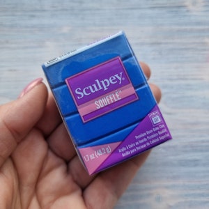 Puede incluir: Un paquete azul de arcilla para hornear Sculpey Souffle. El paquete está etiquetado con el nombre de la marca, el nombre del producto, el peso (1,7 oz o 48,2 g) y las instrucciones para hornear la arcilla. El paquete se sostiene en una mano.