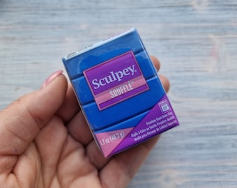 May include: A blue package of Sculpey Souffle brand oven-bake clay. The package is labeled with the brand name, product name, weight (1.7 oz or 48.2 g), and instructions for baking the clay. The package is held in a hand.