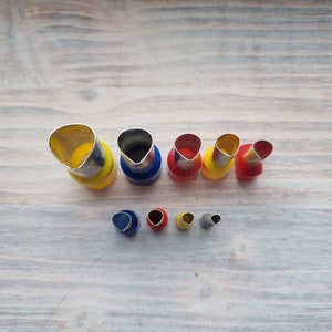 May include: Set of nine metal flower cutters in various sizes. The cutters are yellow, red, and blue. The cutters are arranged in two rows on a wooden surface.