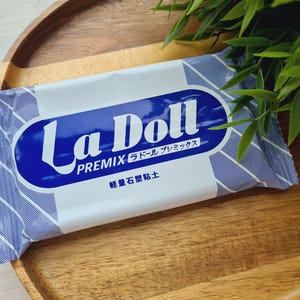 May include: A packet of La Doll Premix clay, with a blue and white design and Japanese text. The packet rests on a wooden tray, with green foliage in the background. The clay is a lightweight stone clay.