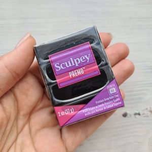 Op de afbeelding: Een zwarte blok Sculpey Premo oven-bakklei van 57 gram (2 ounce). De verpakking heeft een paars en wit label met de merknaam en productinformatie.