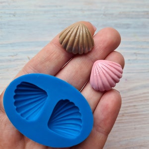 Peut inclure: Un moule en silicone bleu avec deux cavités en forme de coquille. Deux petites coquilles, une brune et une rose, sont présentées à côté du moule.