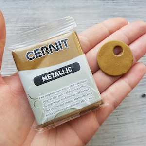 May include: A small package of gold metallic polymer clay by Cernit. The package has text in multiple languages describing the product. The text reads: "Pâte polymère avec micas effet métallique Polymer clay with metallic mica's effect Polymermasse mit Glimmer-Metalleffekt Pasta polimerica con mica effetto metallico Arcilla polimerica metálica con efecto mica"