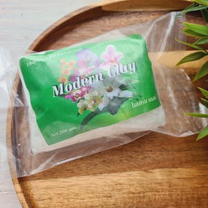 Puede incluir: Un paquete de Modern Clay para manualidades, peso neto 200 g, en un envoltorio de plástico transparente. La etiqueta verde presenta un diseño floral con orquídeas. El paquete está sobre una bandeja de madera con hojas verdes.