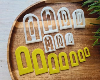 May include: A set of 10 white plastic cookie cutters shaped like arched windows. The cutters are arranged on a wooden surface. There are also 7 yellow cookie cutters shaped like arched windows.