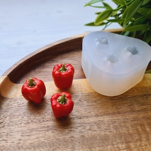 Puede incluir: Tres pimientos rojos en miniatura con tallos verdes están sobre una bandeja de madera junto a un molde de silicona transparente. Los pimientos son de color rojo vibrante. Hay hojas verdes en el fondo.