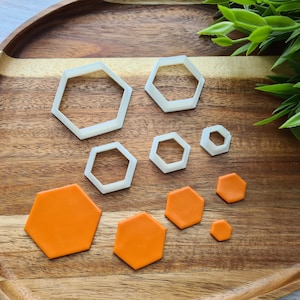 May include: A set of six white plastic hexagon cookie cutters on a wooden surface with six orange hexagon-shaped pieces of fondant.