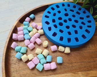 Può includere: Un vassoio di legno contiene una dispersione di marshmallow dai colori pastello e uno stampo in silicone blu brillante, circolare. I marshmallow sono nelle tonalità del rosa, blu e giallo. Lo stampo ha un motivo di fori irregolari.