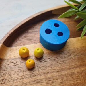 Könnte beinhalten: Eine blaue Silikonform mit drei runden Vertiefungen auf einer Holzoberfläche. Drei kleine gelbe Zitrusfrüchte liegen neben der Form.