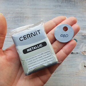 Puede incluir: Un paquete de arcilla polim&eacute;rica met&aacute;lica de la marca Cernit, etiquetado como "Metallic". El paquete est&aacute; en la mano de una persona, junto con un peque&ntilde;o c&iacute;rculo gris con el n&uacute;mero "080" escrito en &eacute;l.