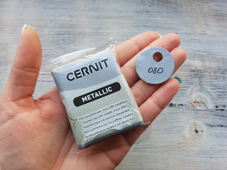 Puede incluir: Un paquete de arcilla polim&eacute;rica met&aacute;lica de la marca Cernit, etiquetado como "Metallic". El paquete est&aacute; en la mano de una persona, junto con un peque&ntilde;o c&iacute;rculo gris con el n&uacute;mero "080" escrito en &eacute;l.