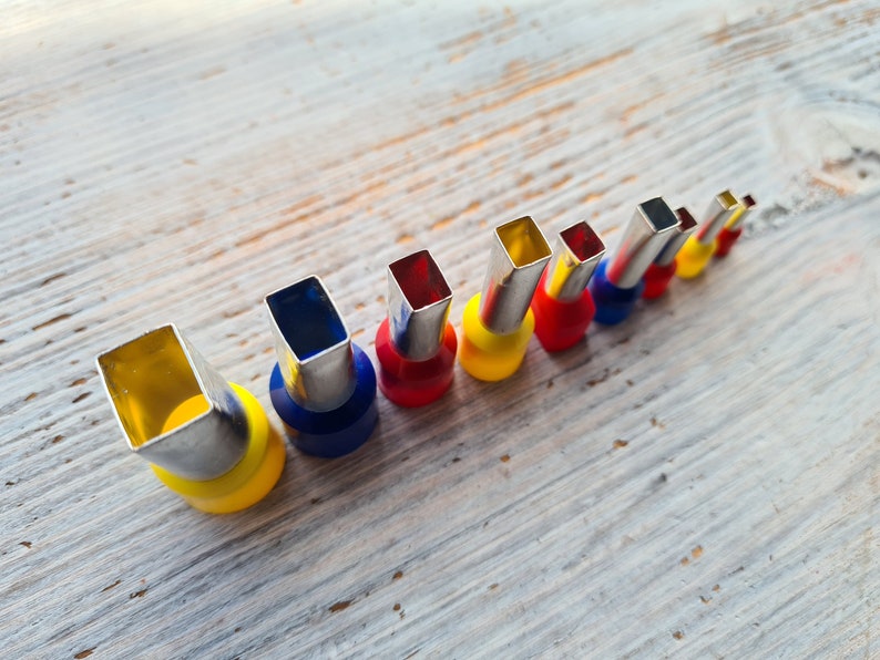 May include: An assortment of electrical wire crimp connectors, arranged in a diagonal line. The connectors have a silver metal square end, with colored plastic bases in yellow, blue, and red. The background is a light-colored wooden surface.