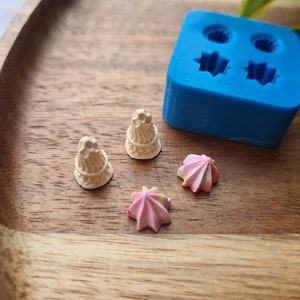 Puede incluir: Cuatro pequeños adornos para pasteles decorativos sobre una superficie de madera. Dos son blancos y con forma de pastel con glaseado, y dos son rosas y con forma de remolino.