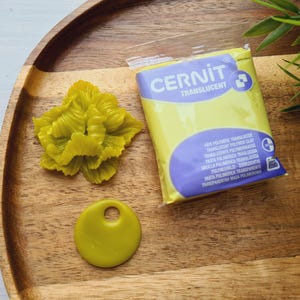 May include: A wooden tray holds a yellow polymer clay flower, a round pendant with a hole, and a package of Cernit translucent polymer clay. The package has a yellow background with a blue label.