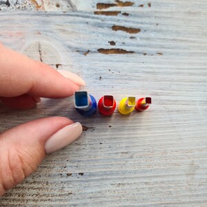May include: A collection of four colorful square-shaped paper punches in varying sizes. The punches have blue, red, and yellow accents. The smallest punch is red, and the largest is blue. The punches are held by a hand.