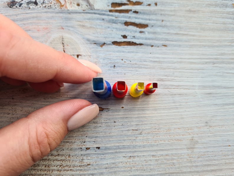 May include: A collection of four colorful square-shaped paper punches in varying sizes. The punches have blue, red, and yellow accents. The smallest punch is red, and the largest is blue. The punches are held by a hand.