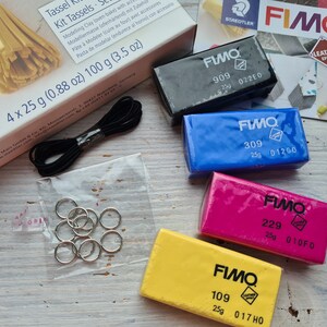 May include: Four blocks of Fimo polymer clay in different colours: black, blue, pink, and yellow. Each block is labelled with the Fimo brand name, a number, and a code. The blocks are on a wooden surface.