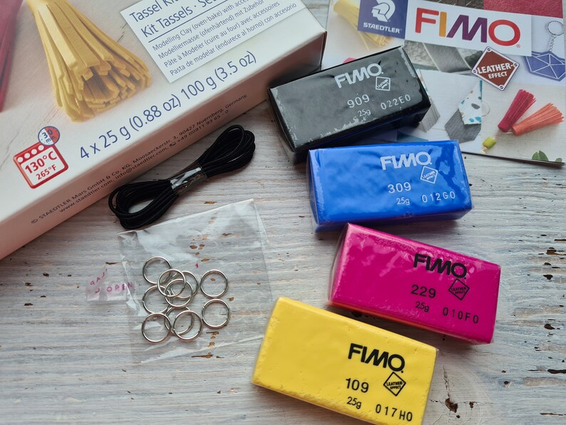 May include: Four blocks of Fimo polymer clay in different colours: black, blue, pink, and yellow. Each block is labelled with the Fimo brand name, a number, and a code. The blocks are on a wooden surface.
