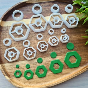 Könnte beinhalten: Ein Set aus weißen Plastik-Ausstechern in verschiedenen Formen, darunter Kreise, Sechsecke und ein dreidimensionales Sechseck-Design. Die Ausstecher sind auf einer Holzoberfläche mit grünen Fondantformen angeordnet.