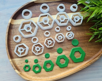 May include: A set of white plastic cookie cutters in various shapes, including circles, hexagons, and a three-dimensional hexagon design. The cutters are arranged on a wooden surface with green fondant shapes.