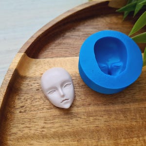 May include: A pale pink face sculpture with a smooth surface, beside a bright blue mould of the same face. The face has closed eyes and a calm expression. The items are on a wooden tray with green foliage.