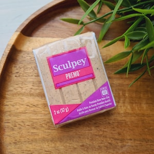 May include: A package of Sculpey Premo oven-bake clay, 57 g, in a beige colour. The packaging has a pink and purple label with the brand name and product details. The clay is on a wooden tray with green leaves.