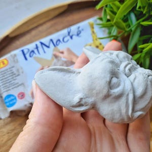 Pode incluir: Uma cabe&ccedil;a de bulldogue franc&ecirc;s esculpida em cinza, feita de papel mach&eacute;. A cabe&ccedil;a est&aacute; sendo segurada em uma m&atilde;o, e o fundo mostra um pacote de papel mach&eacute;.